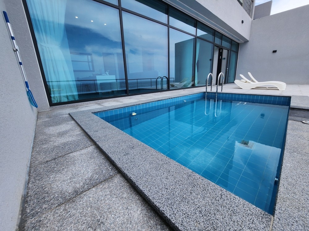 A private plunge pool is shown with clear blue water, bordered by light gray stone tiles. Two lounge chairs are positioned nearby, facing large glass windows that reflect the sky.