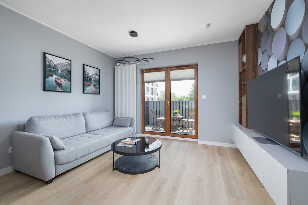 A spacious living room features a fold-out sofa and a circular coffee table. Two framed landscape images adorn the wall, while a TV is mounted on a stylish unit. Sliding glass doors lead to a balcony, enhancing natural light in the room.