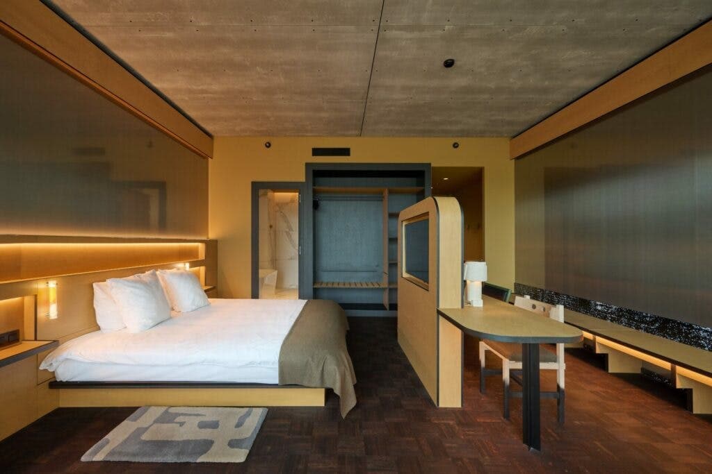 A double room features a spacious bed with crisp white linens, complemented by soft, neutral tones throughout. A small wooden table and chairs are positioned near the window, providing a cozy workspace. Natural light fills the room, highlighting the minimalist design and warm atmosphere.