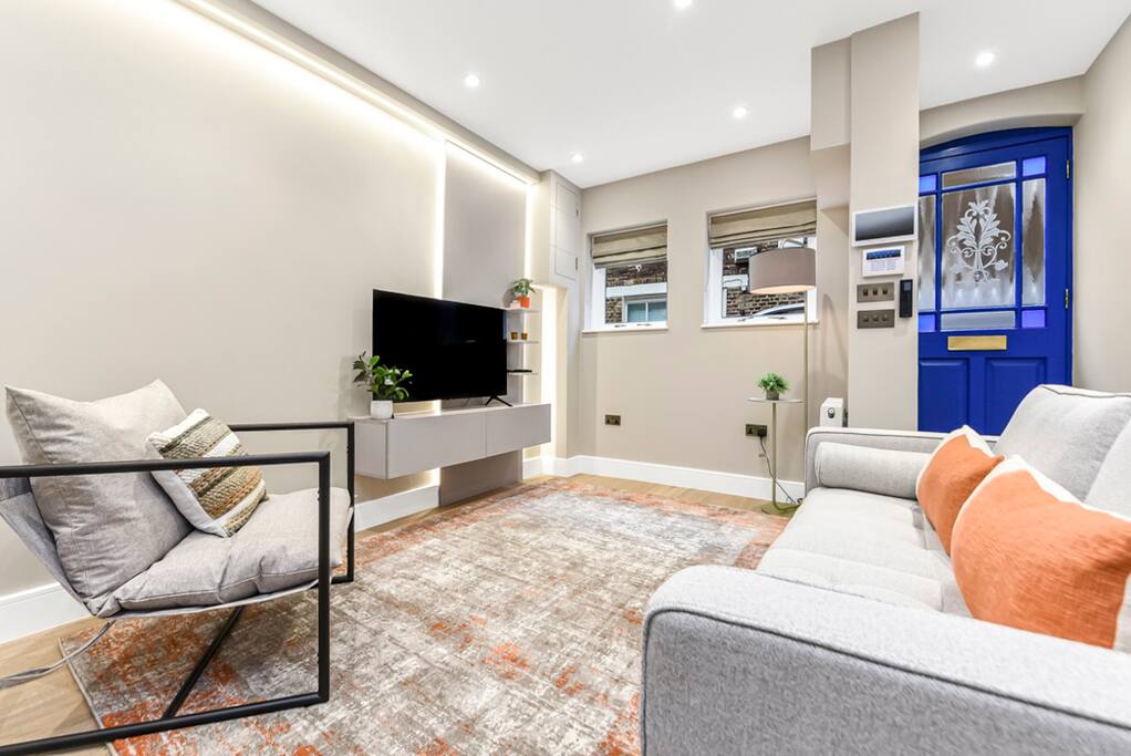 A modern living space is illuminated with soft, warm lighting. A sleek gray sofa and a patterned armchair provide comfortable seating. A flat-screen TV is mounted on the wall opposite a console table, while a vibrant area rug adds texture to the floor. The entry door features a bright blue hue.