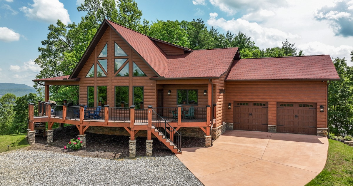 A spacious luxury cabin is showcased with a wooden exterior, featuring large windows that offer scenic valley views. A welcoming porch extends from the front, leading down to a driveway beside a two-car garage. Lush greenery surrounds the property, enhancing the natural setting.