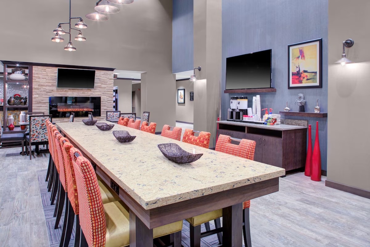 A spacious dining area is highlighted by a long stone table surrounded by tall chairs with orange upholstery. The room is accented by modern lighting fixtures and features a decorative bar area with a coffee machine and varied decor elements.