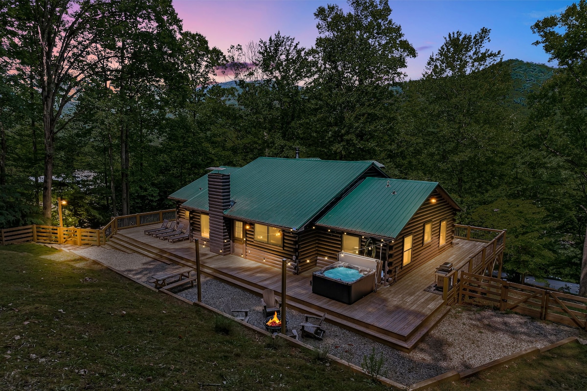 A two-story log cabin is surrounded by lush trees, with a spacious wrap-around deck featuring a hot tub and seating areas. A fire pit is positioned on the lower level of the deck, illuminated by soft outdoor lighting against the twilight sky.