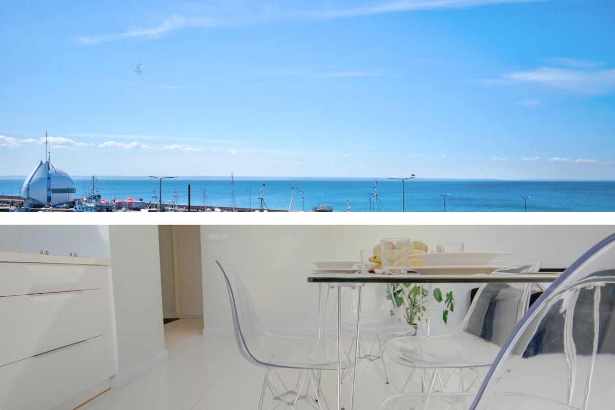 A bright dining area features a clear table surrounded by transparent chairs. In the background, large windows frame a view of the expansive blue sky and calm sea, reflecting light and creating an airy atmosphere.
