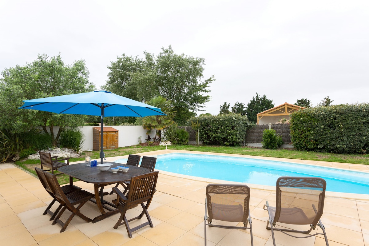 A spacious terrace is highlighted with a wooden dining table set beneath a large blue umbrella. Several lounge chairs are arranged nearby, facing a private outdoor swimming pool surrounded by greenery. A fence and additional landscaping provide privacy and a serene ambiance.