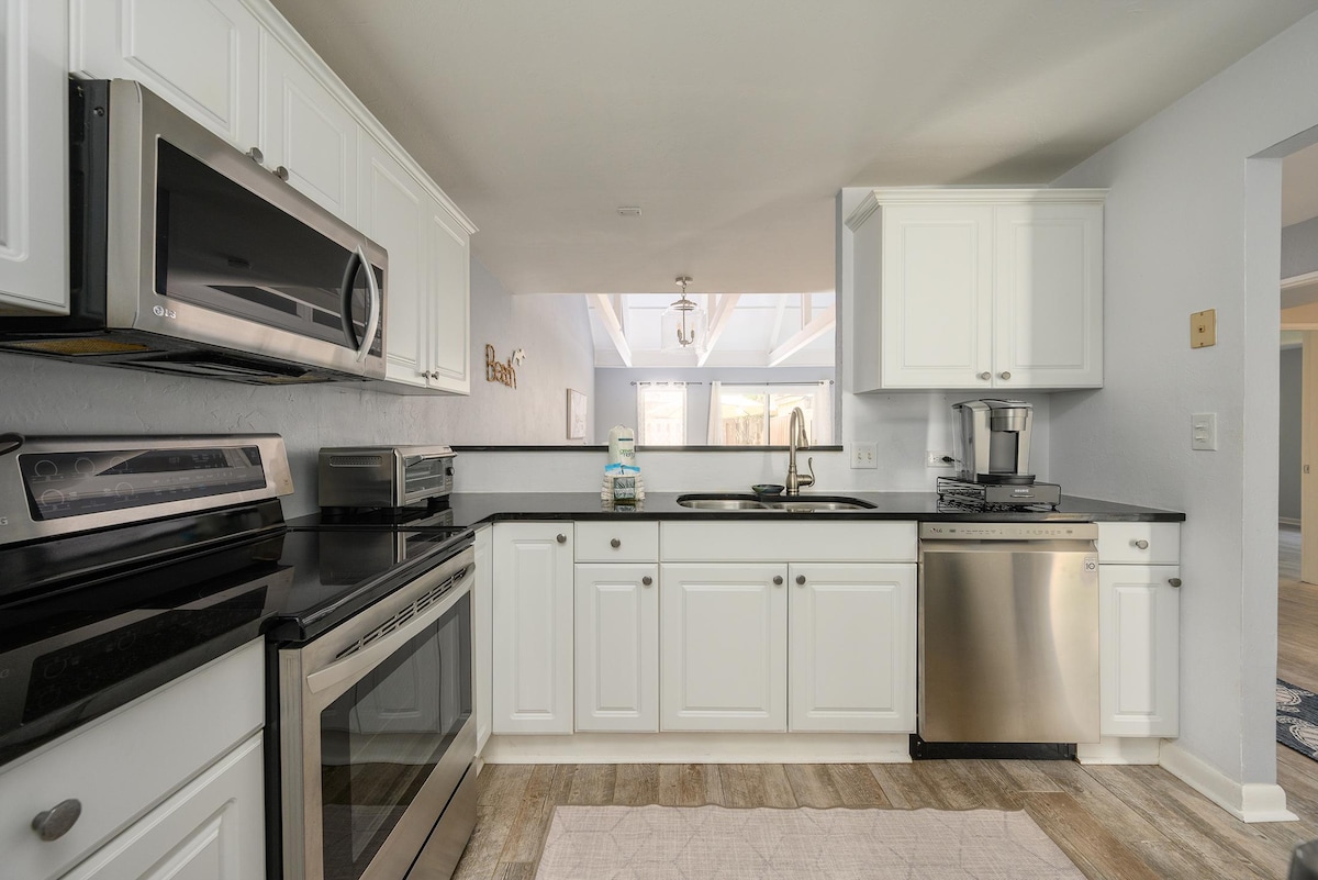 The kitchen is equipped with stainless steel appliances, including a microwave, oven, and dishwasher. White cabinetry contrasts with a dark countertop, creating a clean, modern look. A window above the sink allows natural light to brighten the space, enhancing the open layout.