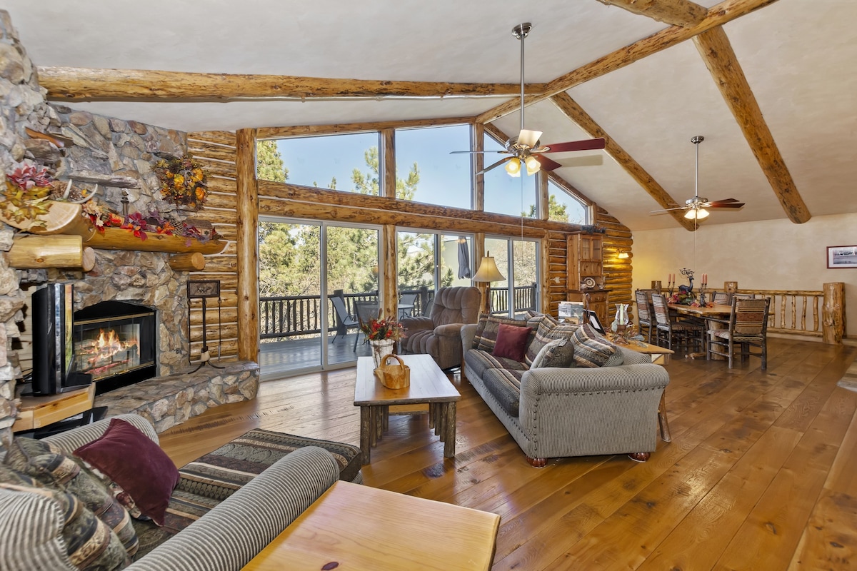The living area features rustic wooden beams and a stone fireplace, creating a cozy focal point. Large windows offer ample natural light and forest views. Comfortable seating arrangements accommodate guests, while a dining area is visible, enhancing the inviting atmosphere for gatherings.