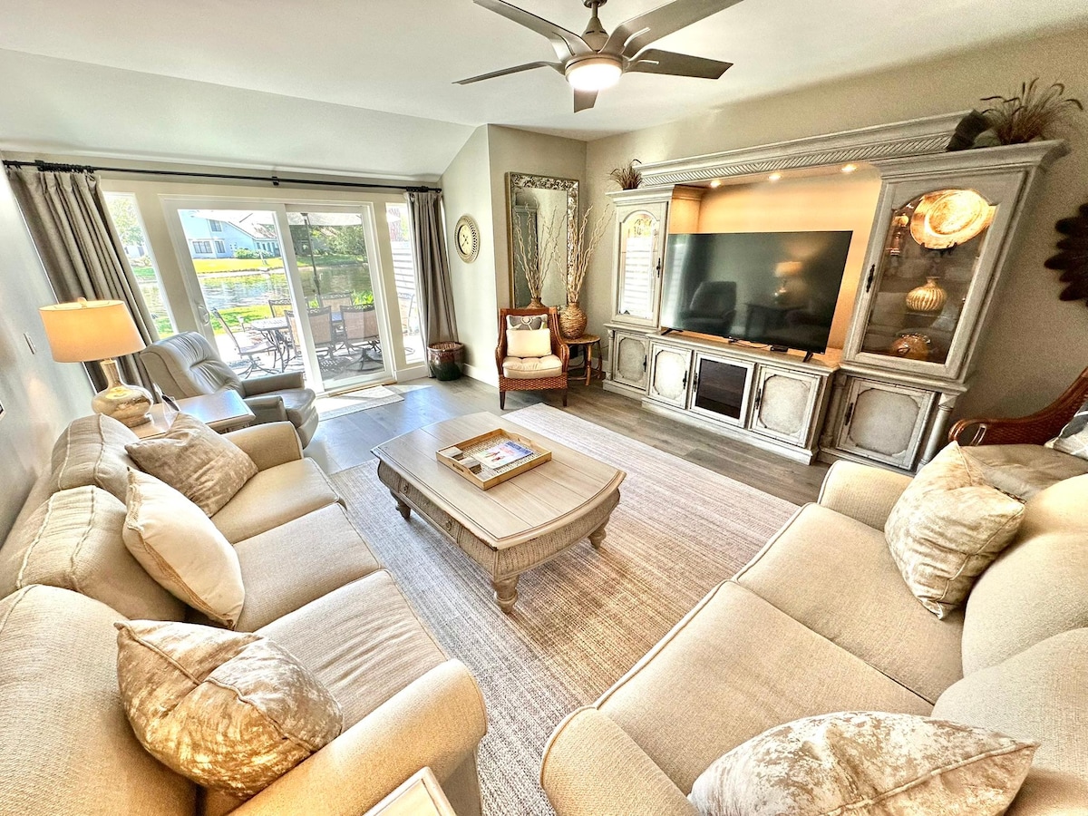 A spacious living area features two light-colored sofas and an armchair, arranged around a central coffee table. A large flat-screen television is mounted on an elegant cabinet. Sunlight filters through sliding glass doors, leading to a patio with views of the serene lake.
