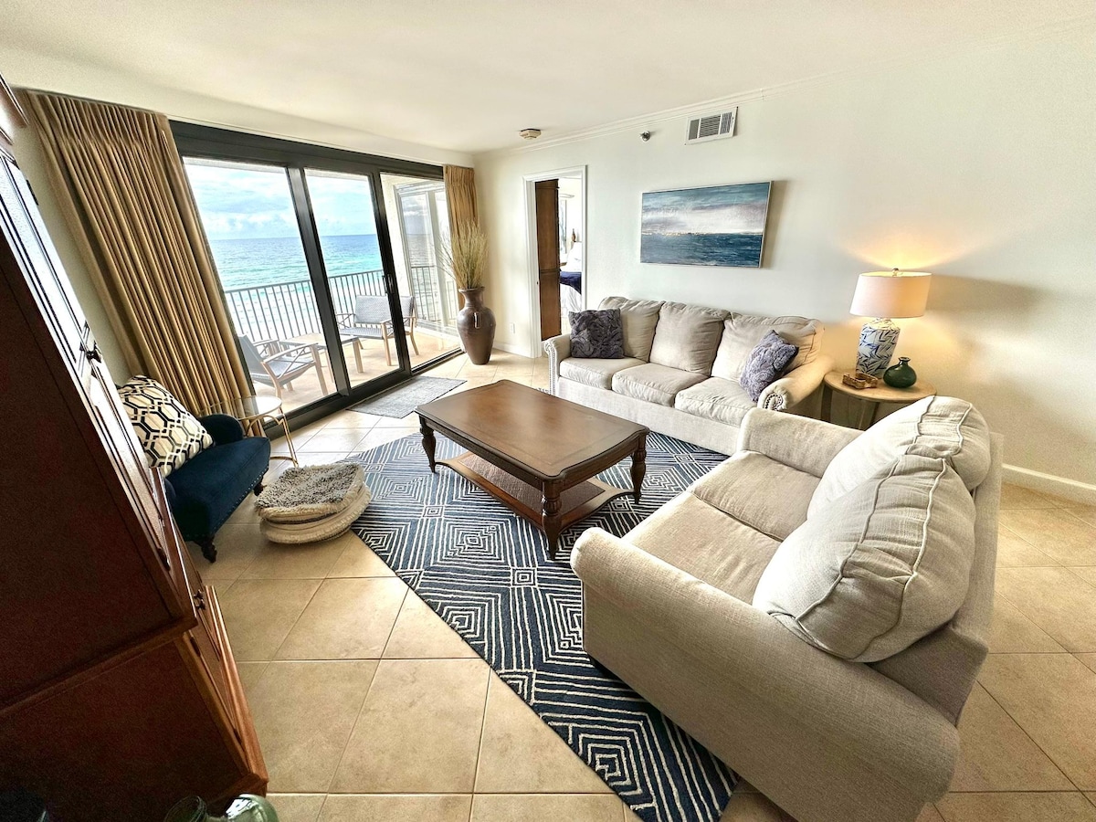 The living area features two comfortable sofas facing a coffee table, with a patterned area rug beneath. Floor-to-ceiling glass doors offer views of the beach and lead to the private balcony, while soft natural light illuminates the space.