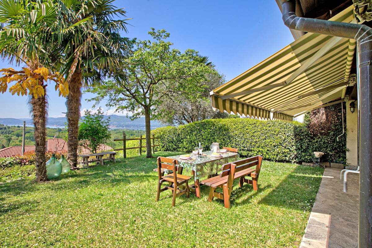 A private garden area features a table set with chairs, surrounded by lush greenery and palm trees. A covered terrace provides shade, enhancing the outdoor dining experience. Scenic views of the hillside can be observed in the background, creating a tranquil setting.