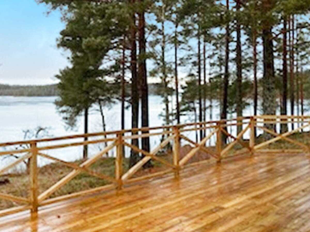 A spacious wooden terrace is visible, bordered by a natural wooden railing. The terrace faces a tranquil lake, surrounded by tall trees, creating a serene outdoor space ideal for relaxation.