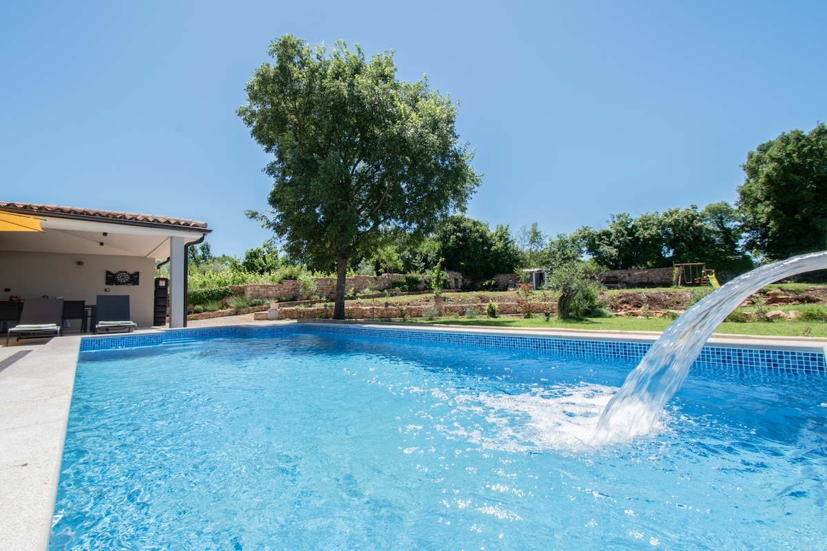 A clear blue pool features a waterfall and is surrounded by lush greenery. Sun loungers and an umbrella can be seen nearby, providing a space for relaxation. The inviting outdoor area also includes spaces for leisure activities in the backdrop.
