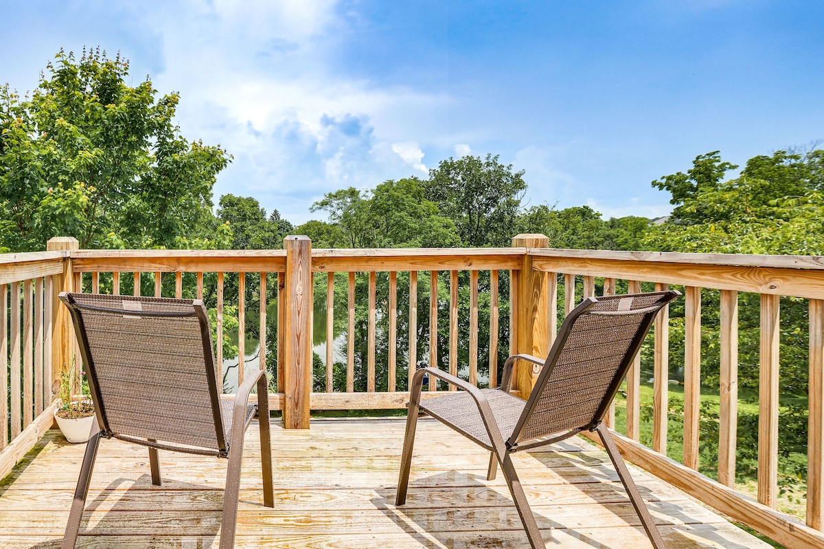 A private deck overlooks lush greenery, featuring two outdoor chairs positioned beside a low railing. The wooden surface is inviting, and the blue sky above complements the serene outdoor setting.