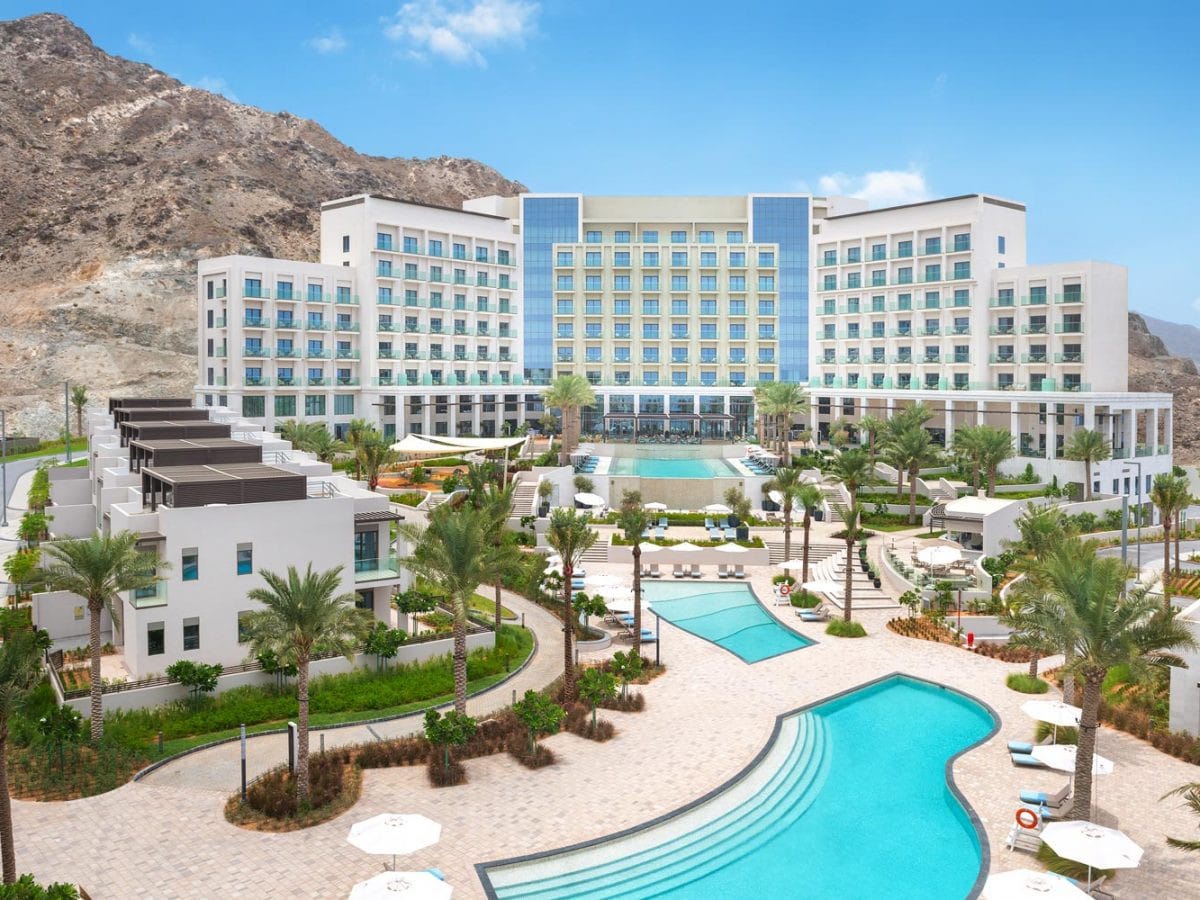 A modern beachfront resort is surrounded by lush landscaping and palm trees. Multiple swimming pools are visible, with a spacious terrace leading to the main building. The structure features large glass windows and a mix of contemporary architecture against a backdrop of mountains.