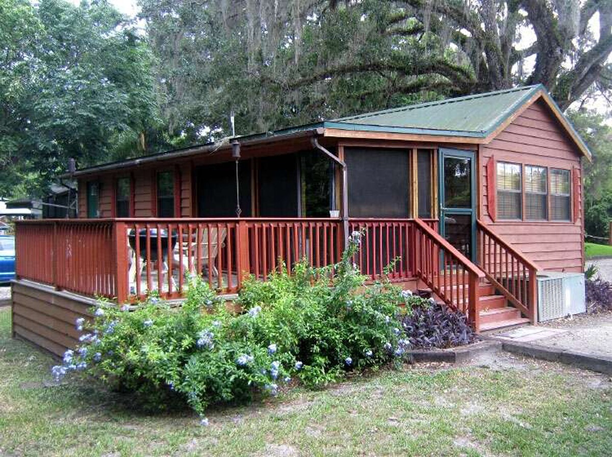 A cabin features rustic cedar siding and a welcoming porch surrounded by lush greenery. A well-maintained garden area with flowering plants is situated in front, while large windows allow for natural light to enter the cozy interior.