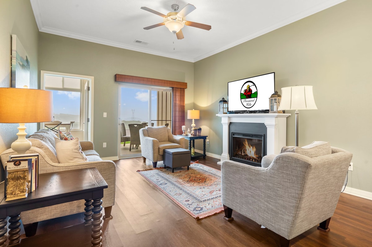 A spacious living area is highlighted by a seasonal fireplace and features comfortable seating, including two armchairs and a large sofa. A coffee table rests in the center, while a flat-screen TV is mounted above the fireplace. Natural light enters through the sliding glass door leading to the outside.