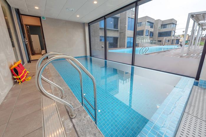 An indoor pool area is shown, featuring a staircase for entry into the clear blue water. Large windows provide natural light, while a lifebuoy rests against the wall. The pool is surrounded by a neutral-toned tiled floor and offers views of the outdoor spaces.
