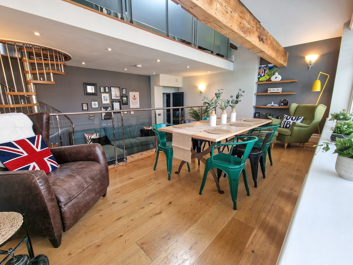 An open-plan living area features a large wooden dining table surrounded by green chairs. A cozy brown leather armchair with a Union Jack cushion adds character, while a modern spiral staircase leads to an upper level. Warm light is provided by wall sconces against a dark grey backdrop.