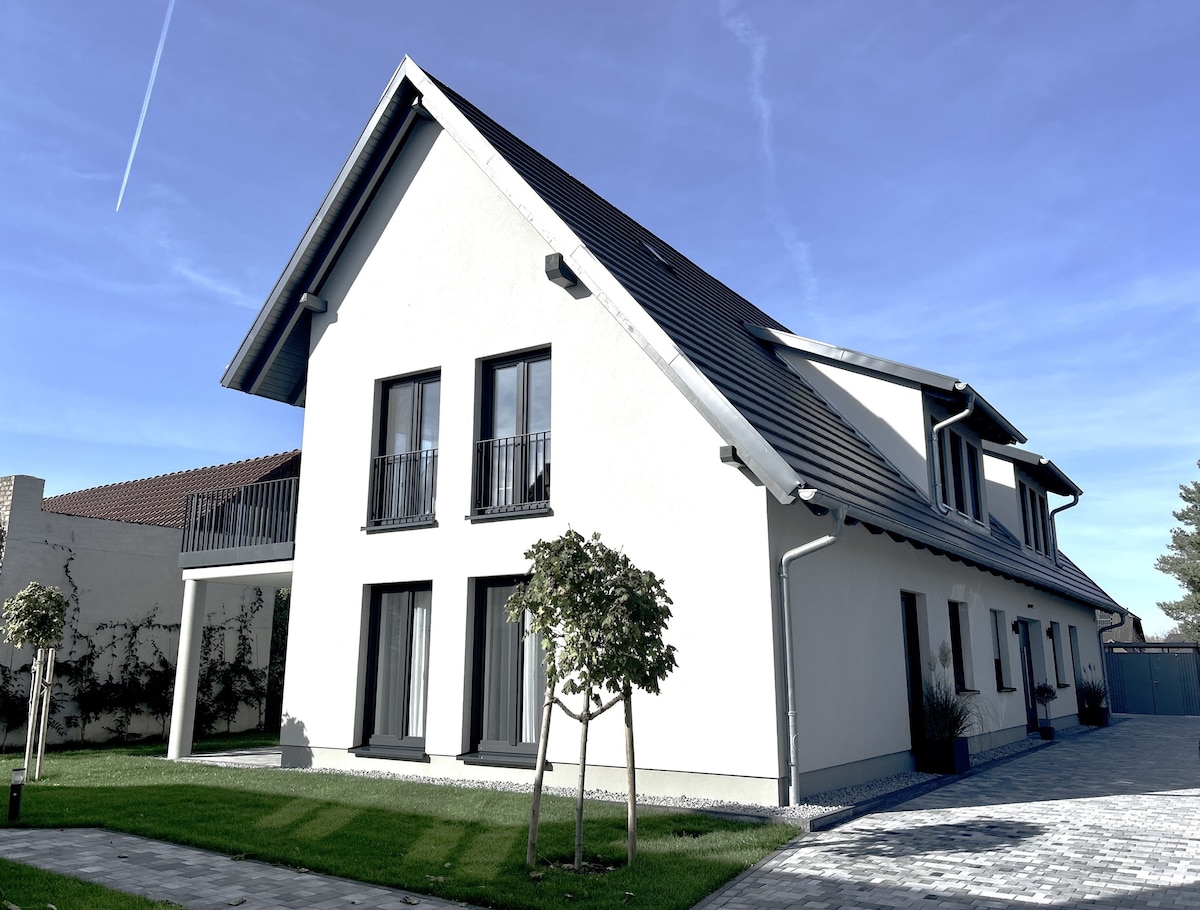 The modern flat complex is presented with a clean white facade and a sloping roof. Balconies are visible on the upper level, framed by well-maintained greenery. A paved pathway leads to the entrance, enhancing the inviting exterior in natural sunlight.
