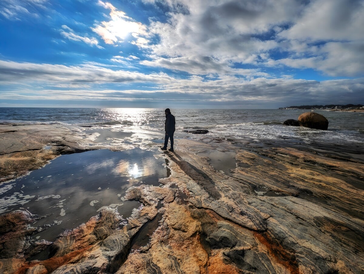 A figure stands on a rocky shoreline, with the ocean stretching out in the background. Reflections of the sky and clouds shimmer on the wet rocks, while waves gently lap at the edges. The landscape is accented by a mix of earthy tones and sandy hues.