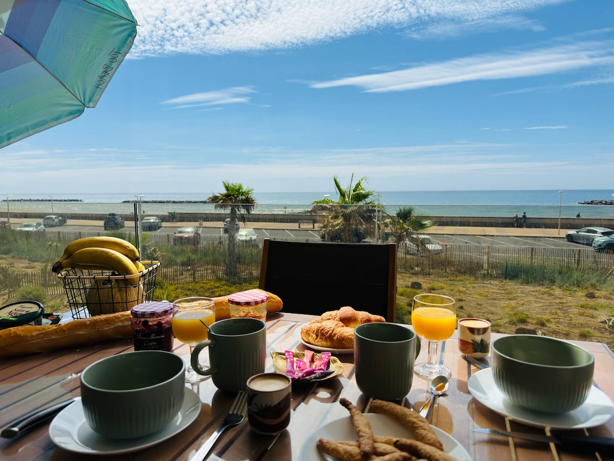 A breakfast table is set with plates, cups, and beverages, overlooking a calm sea view. The table features an array of breakfast items, including croissants, fruit, and jams. The backdrop includes a bright sky and palm trees, creating a peaceful atmosphere.