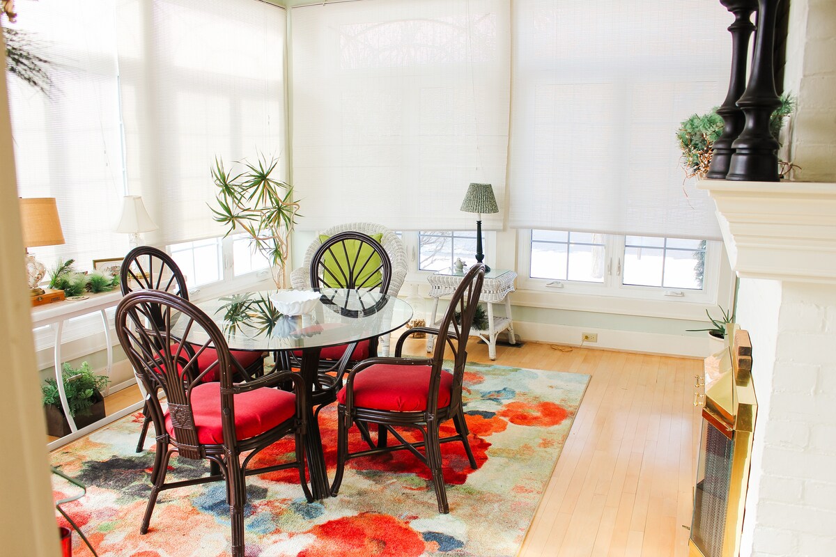 A bright sunroom features a round glass table surrounded by four chairs with red upholstery. A colorful floral area rug lies beneath the table, and large windows allow natural light to fill the space. Indoor plants are arranged thoughtfully around the room.