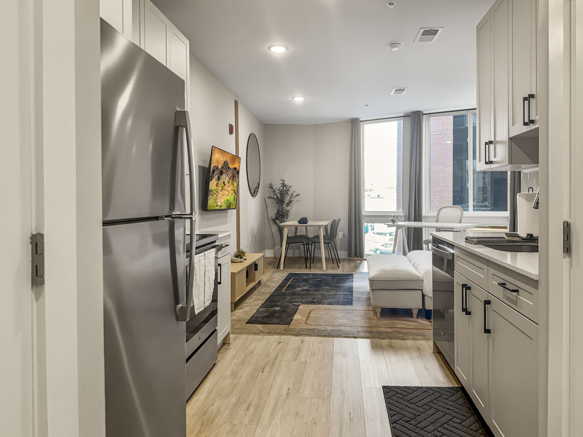 A modern kitchen is visible, featuring stainless steel appliances and a spacious countertop. The open layout leads to a cozy living space with a comfortable couch, dining table, and large window allowing natural light. Neutral tones and contemporary decor create a welcoming ambiance.