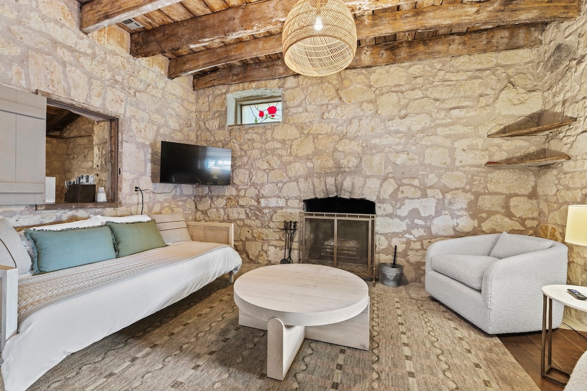 A cozy sitting room is highlighted by rustic stone walls and exposed wooden beams. A plush sofa and modern armchair provide seating, while a round coffee table sits in the center. A seasonal-use fireplace is positioned against the wall, and a TV is mounted nearby.