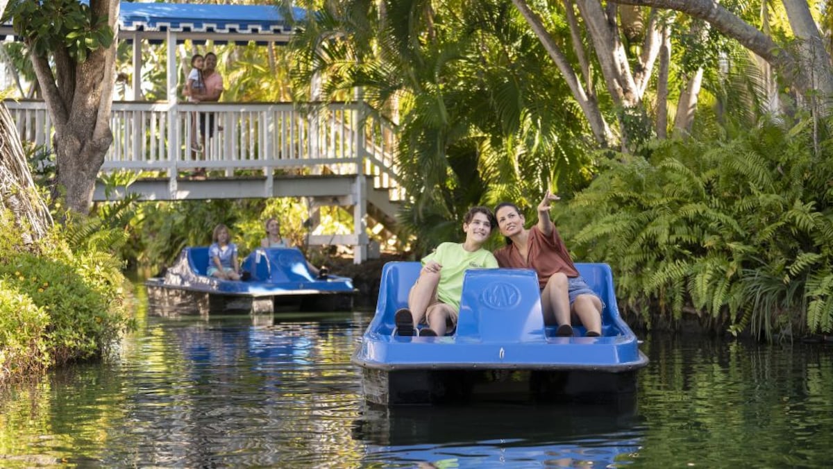 Guests can be seen enjoying a paddleboat ride on a tranquil waterway, surrounded by lush greenery and tropical foliage. A wooden deck leads to a charming structure in the background, adding to the serene atmosphere of the resort.