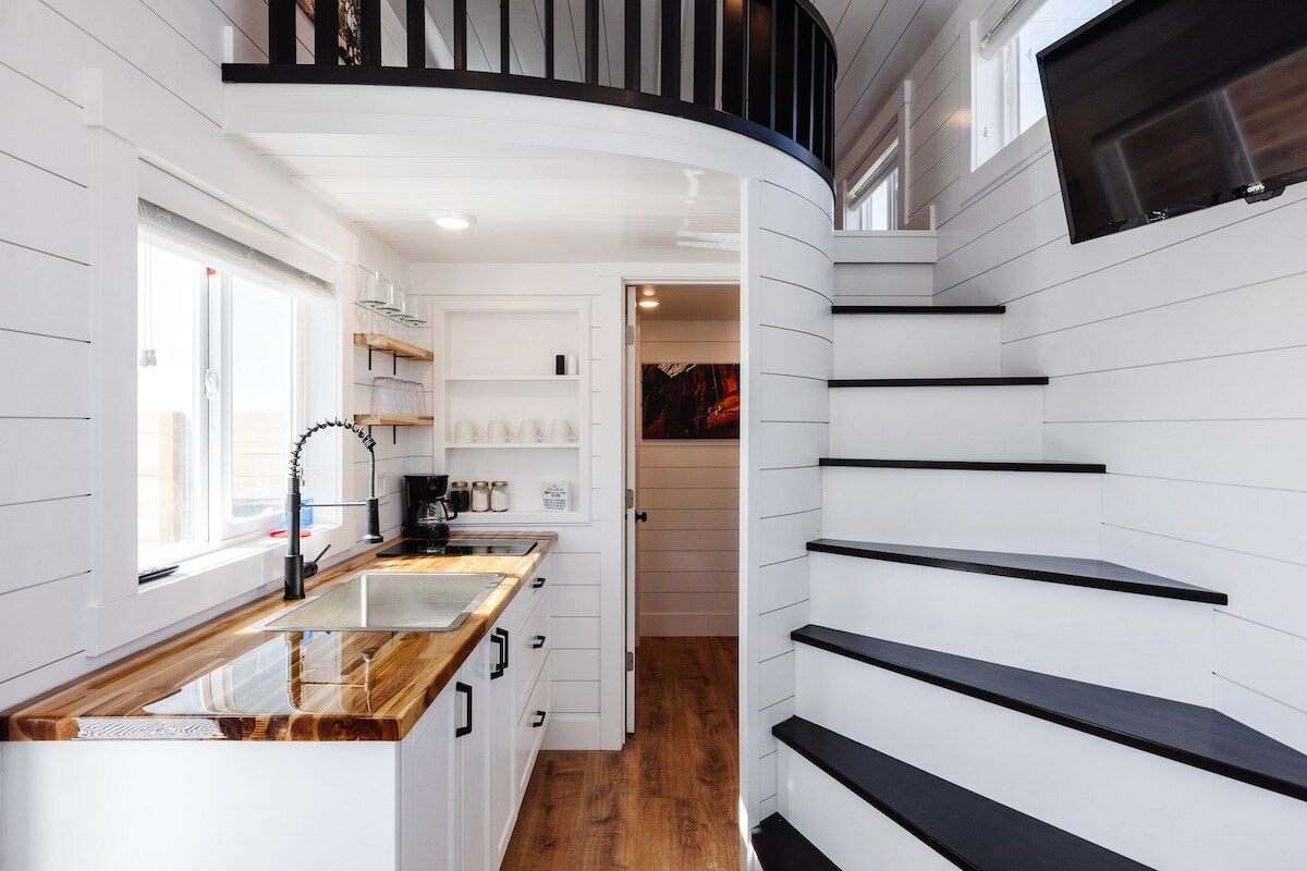 A compact kitchen area features a polished wooden countertop with a stainless steel sink, surrounded by white cabinetry. A staircase with black treads leads to an upper level, while natural light enters through multiple windows.