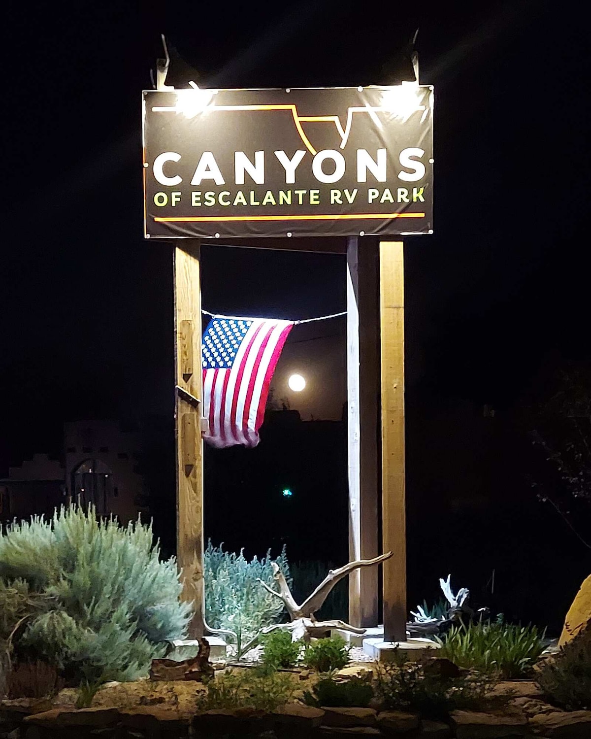 A wooden sign with the text 'Canyons of Escalante RV Park' is illuminated at night, featuring an American flag hanging below it. In the background, a full moon is visible in the night sky, enhancing the serene atmosphere of the park.