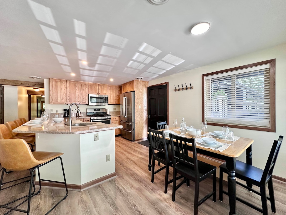 A spacious kitchen and dining area features modern appliances and a large table set for meals. The kitchen includes wooden cabinetry, a stainless steel refrigerator, and bar seating. Light filters through the windows, creating a bright and open environment.