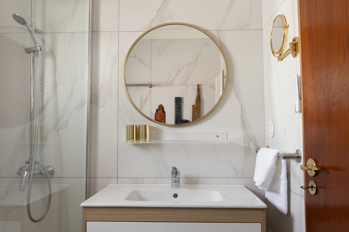 A modern bathroom features elegant marble walls and a round mirror framed in gold. A sleek, light wood vanity with a minimalist sink is positioned below the mirror. A glass shower enclosure is visible in the background, while neatly arranged toiletries are displayed on the shelf.