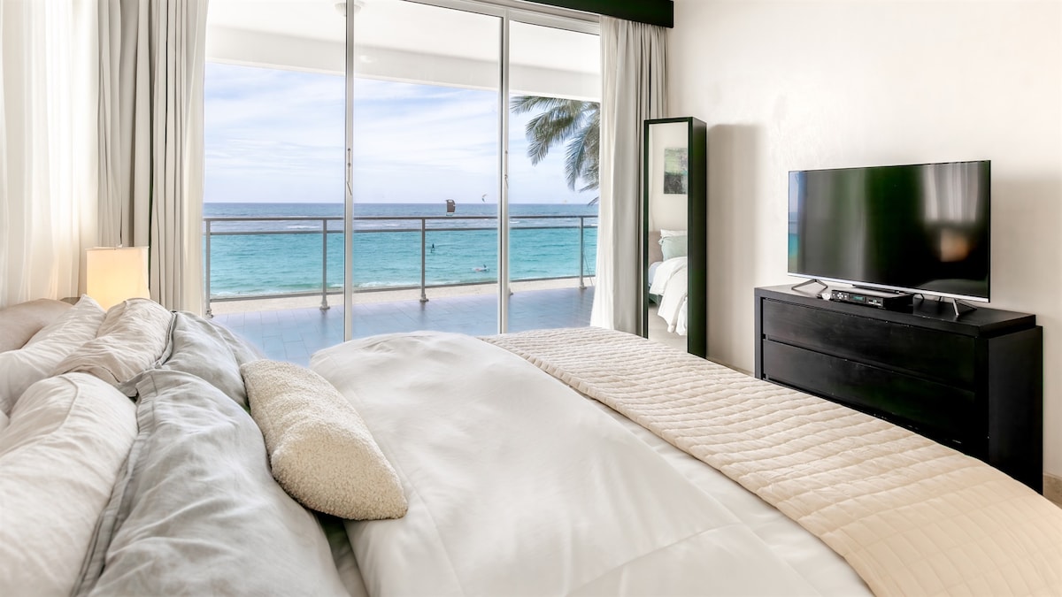 A serene bedroom features a spacious bed with neutral-toned bedding. Large sliding glass doors provide views of the ocean and fill the room with natural light. A flat-screen TV is positioned on a dark wooden dresser, complementing the elegant, minimalistic decor.