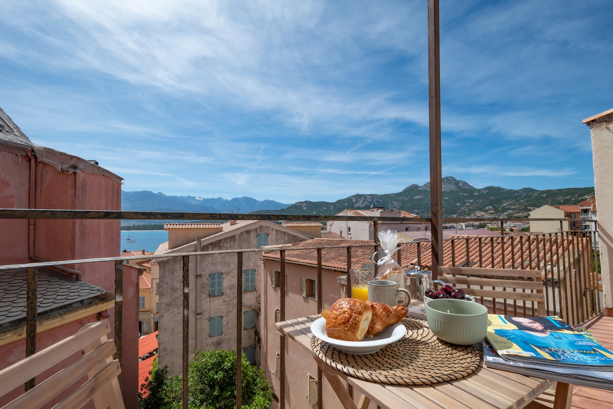 A balcony table is set with breakfast items, including croissants, fruit, and juice. A view of the sea and mountains is visible in the background, while the charming rooftops of Calvi add to the picturesque setting, under a clear blue sky.