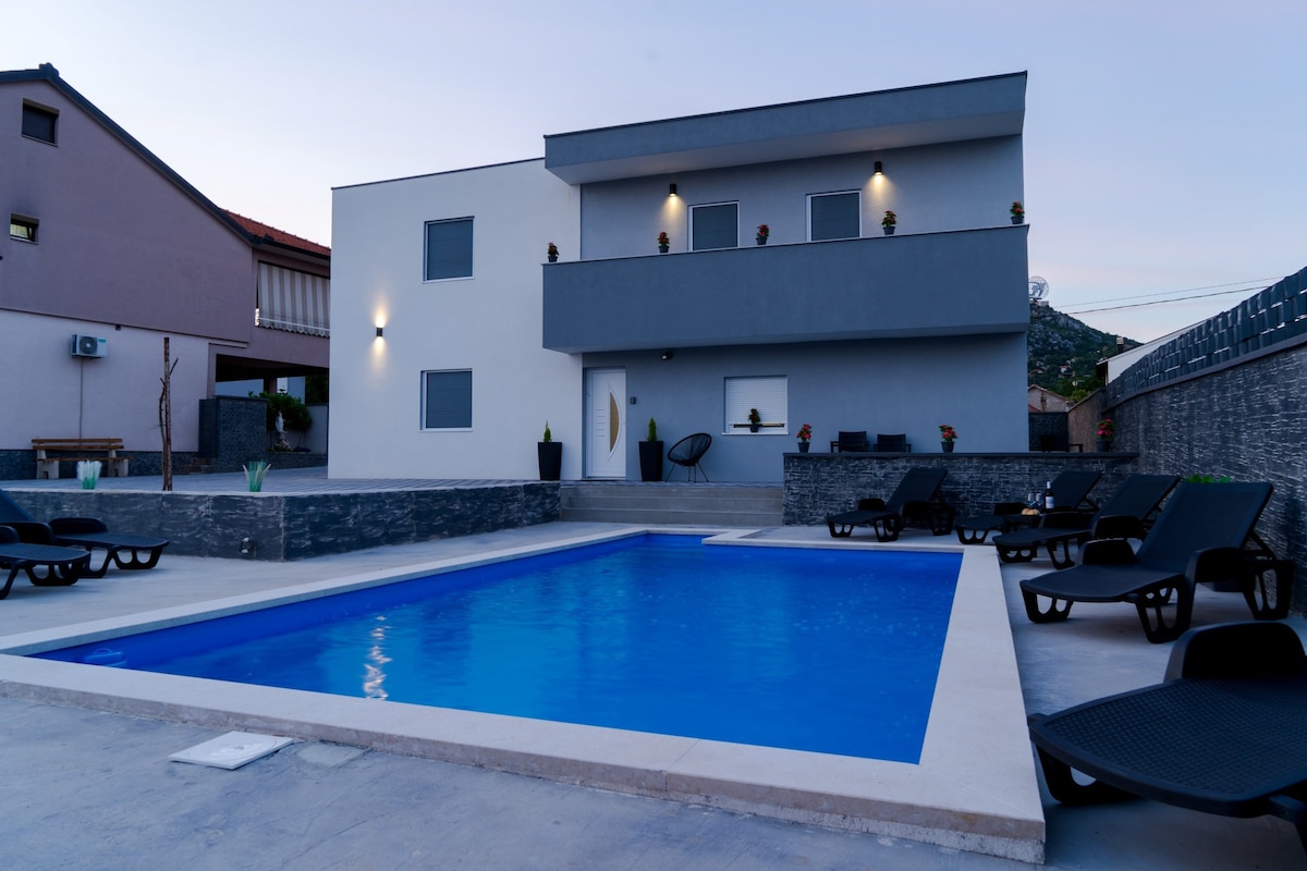A modern, two-story building is depicted beside a clear swimming pool. Lounge chairs surround the pool area, with plants strategically placed along the perimeter. The structure features large windows and simple lines, contributing to an open and welcoming environment.