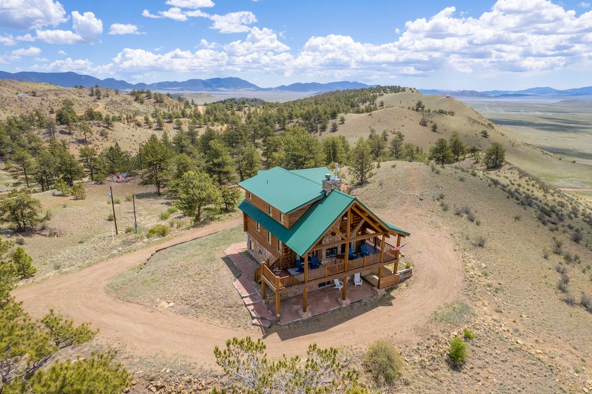 The two-story cabin is surrounded by a 35-acre yard, showcasing rolling hills and distant mountains. A covered deck wraps around the home, offering shaded outdoor space. The unique green metal roof complements the natural landscape under a bright blue sky dotted with clouds.