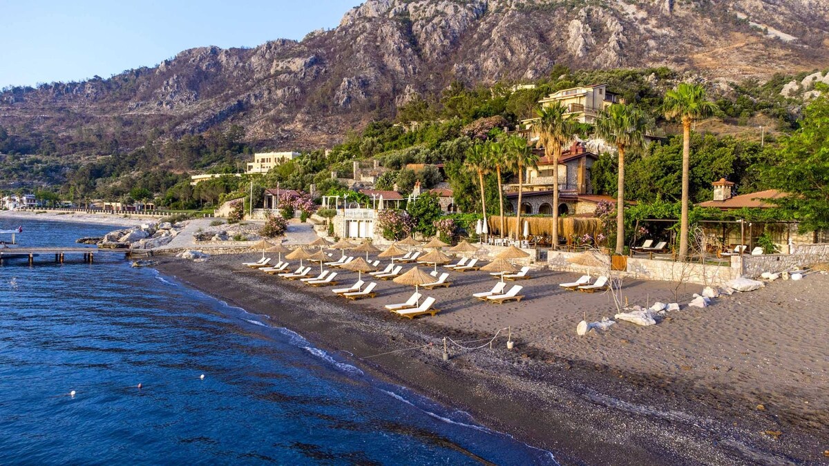 A serene beach area features soft sand and clear waters, lined with sun loungers and parasols. The backdrop includes lush greenery and gently sloping hills, while the tranquil sea reflects the sunlight, creating a peaceful coastal retreat.