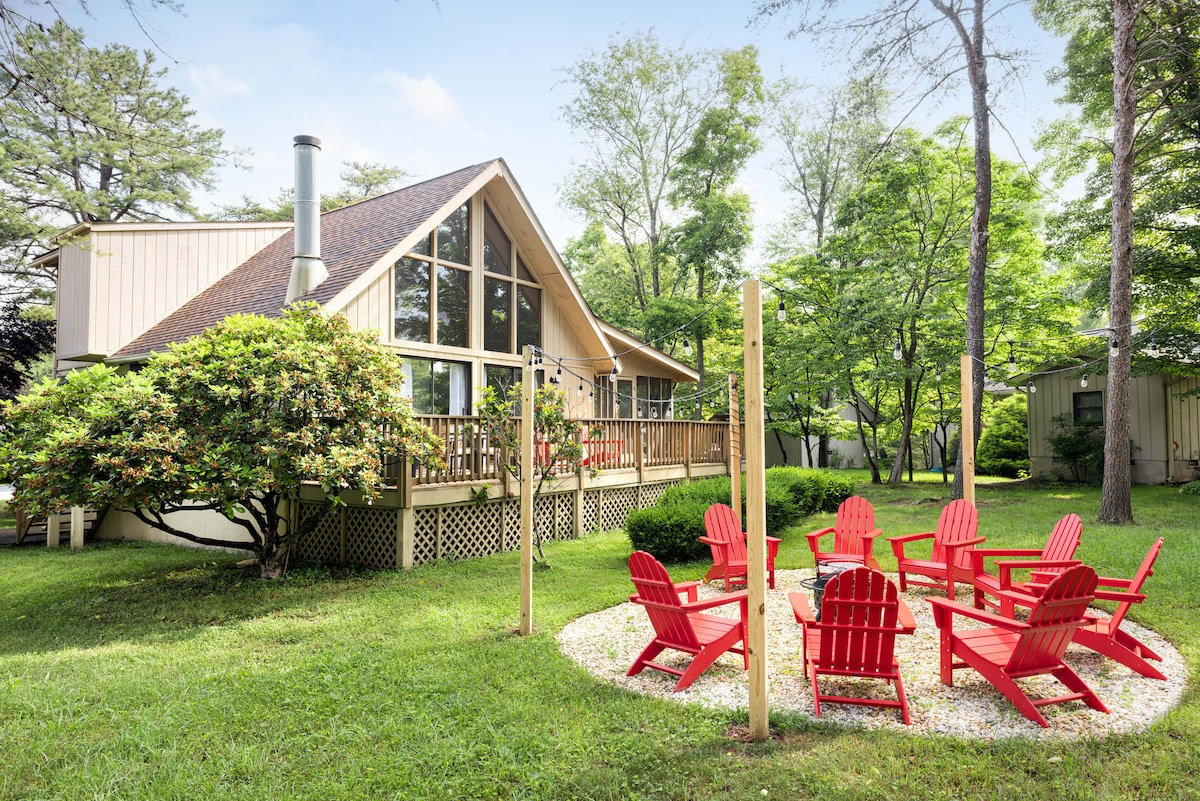 An updated chalet is surrounded by lush greenery, featuring a large deck with ample seating. A fire pit with multiple red Adirondack chairs is situated nearby on a manicured lawn, inviting gatherings and relaxation amidst the serene landscape.
