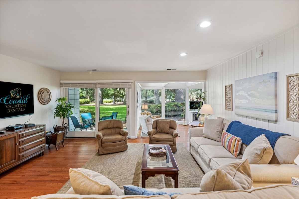 A cozy living area features a light-colored sofa and armchairs arranged around a coffee table, with a flat-screen TV mounted on the wall. Large sliding glass doors provide views of the outdoor space, while soft lighting creates an inviting ambiance.