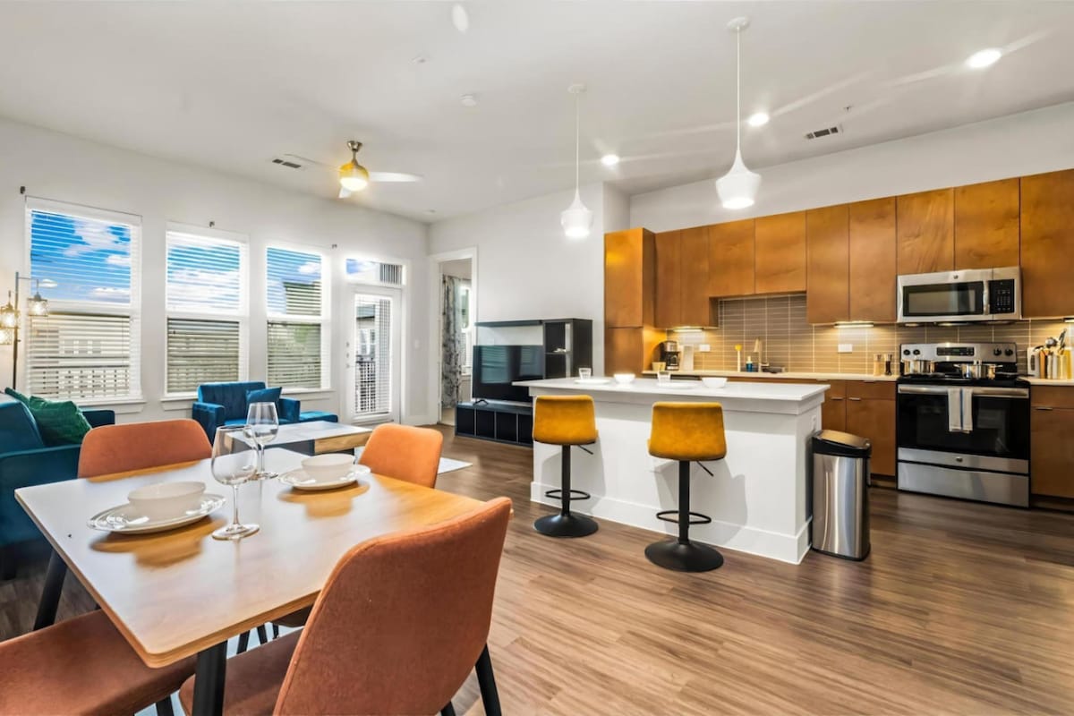 An open concept kitchen and dining area features warm wood cabinetry and modern appliances. A dining table for four is set beside stylish orange chairs. The living space offers a comfortable seating arrangement and large windows that invite natural light, enhancing the overall ambiance.