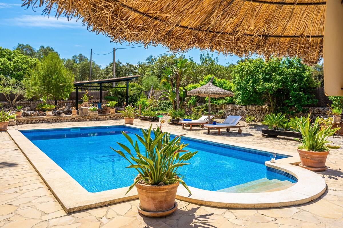 A serene pool area features a clear blue pool surrounded by stone paving and lush Mediterranean vegetation. Comfortable lounge chairs are positioned nearby, and a shaded thatched roof provides relief from the sun. Planters with vibrant green foliage enhance the tranquil setting.