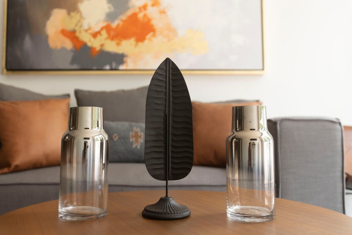 A central coffee table displays two glass vases and a decorative leaf sculpture. The background features a muted palette with an artwork hanging above a comfortable gray couch adorned with warm-toned cushions.