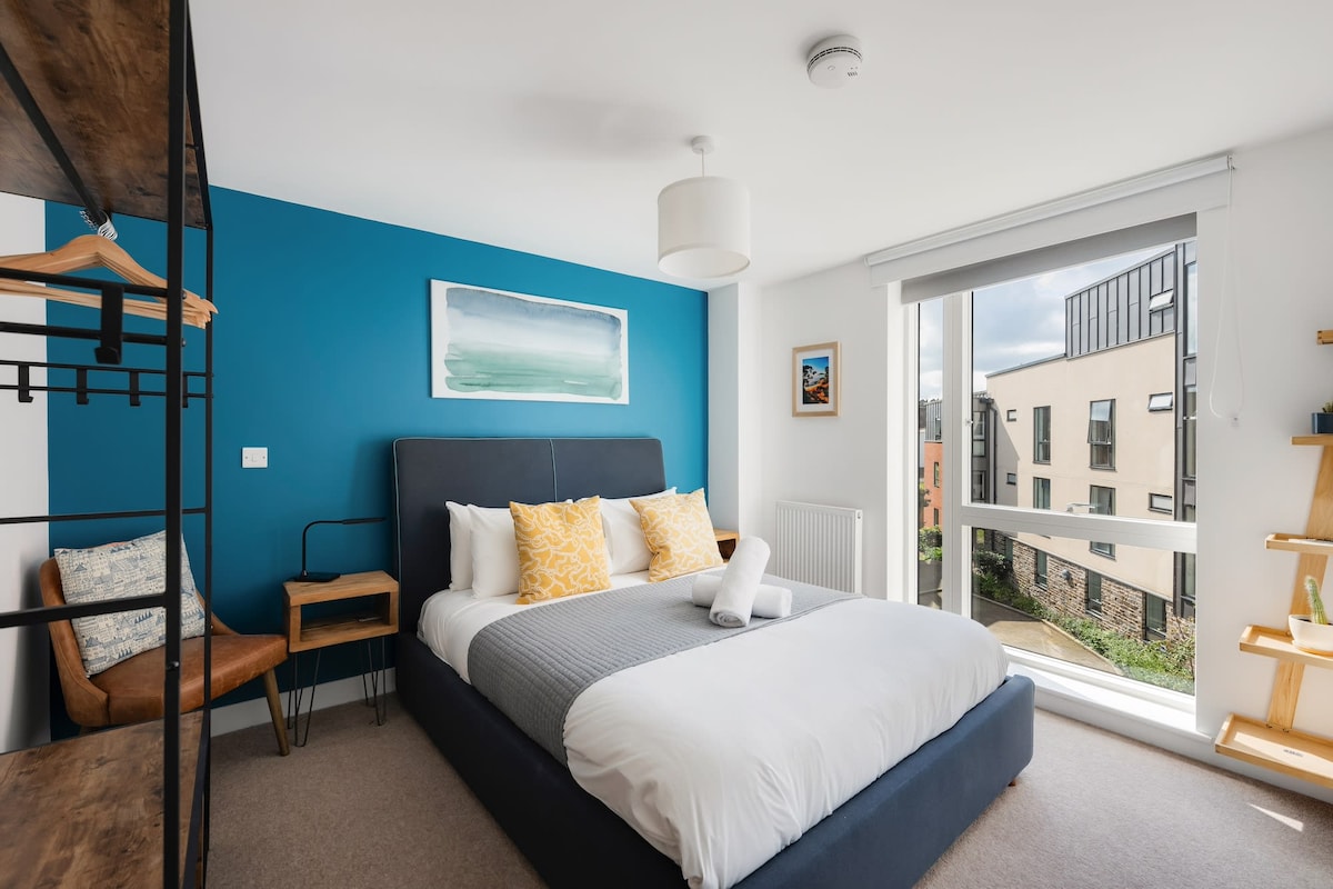 A modern bedroom features a spacious bed with a light duvet and decorative pillows. A blue accent wall complements light wooden shelves and a cozy chair. Natural light floods the room through large windows, offering views of the surrounding buildings.