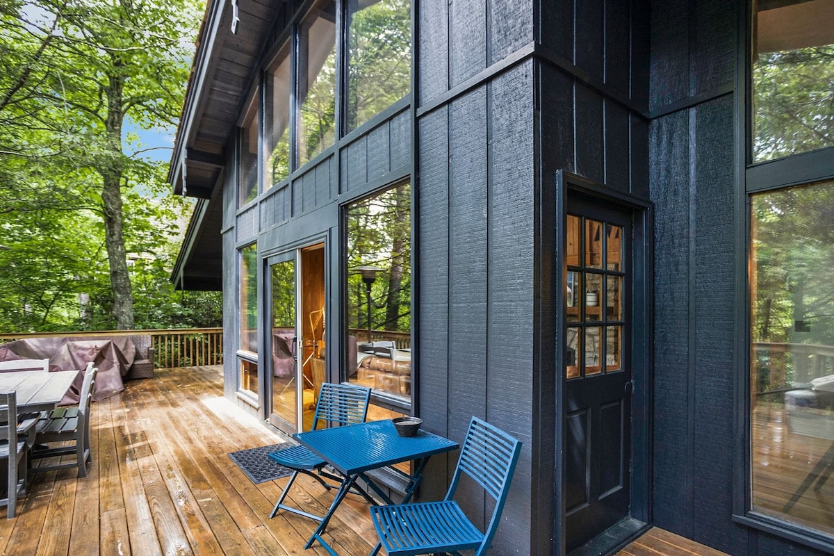 A spacious wooden deck is surrounded by lush greenery, featuring a small blue table and two chairs. Large windows provide views into the home, while the dark exterior contrasts with the natural setting. The entrance door is framed by multiple window panes.