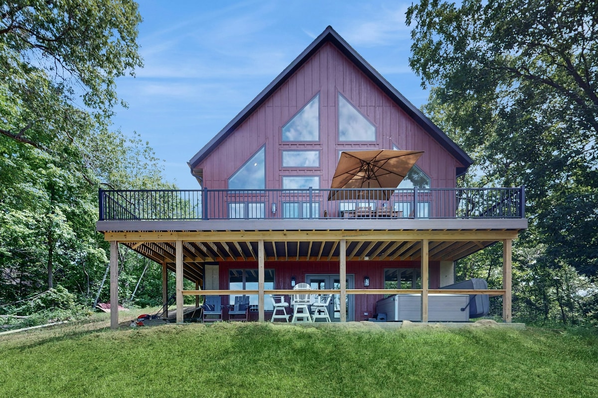 A modern house presents a large deck with a shaded dining area beneath an umbrella. Expansive glass windows highlight the upper level, while lush greenery surrounds the structure. Below, a hot tub is visible, enhancing the outdoor relaxation space.