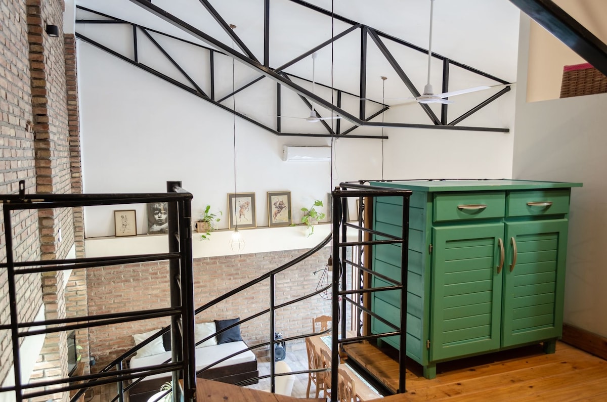 The interior features a spiral staircase leading to a loft area, characterized by high ceilings and exposed beams. A green cabinet is positioned near the railing, adding a pop of color. Framed artwork and potted plants can be seen in the background, contributing to the inviting ambiance.