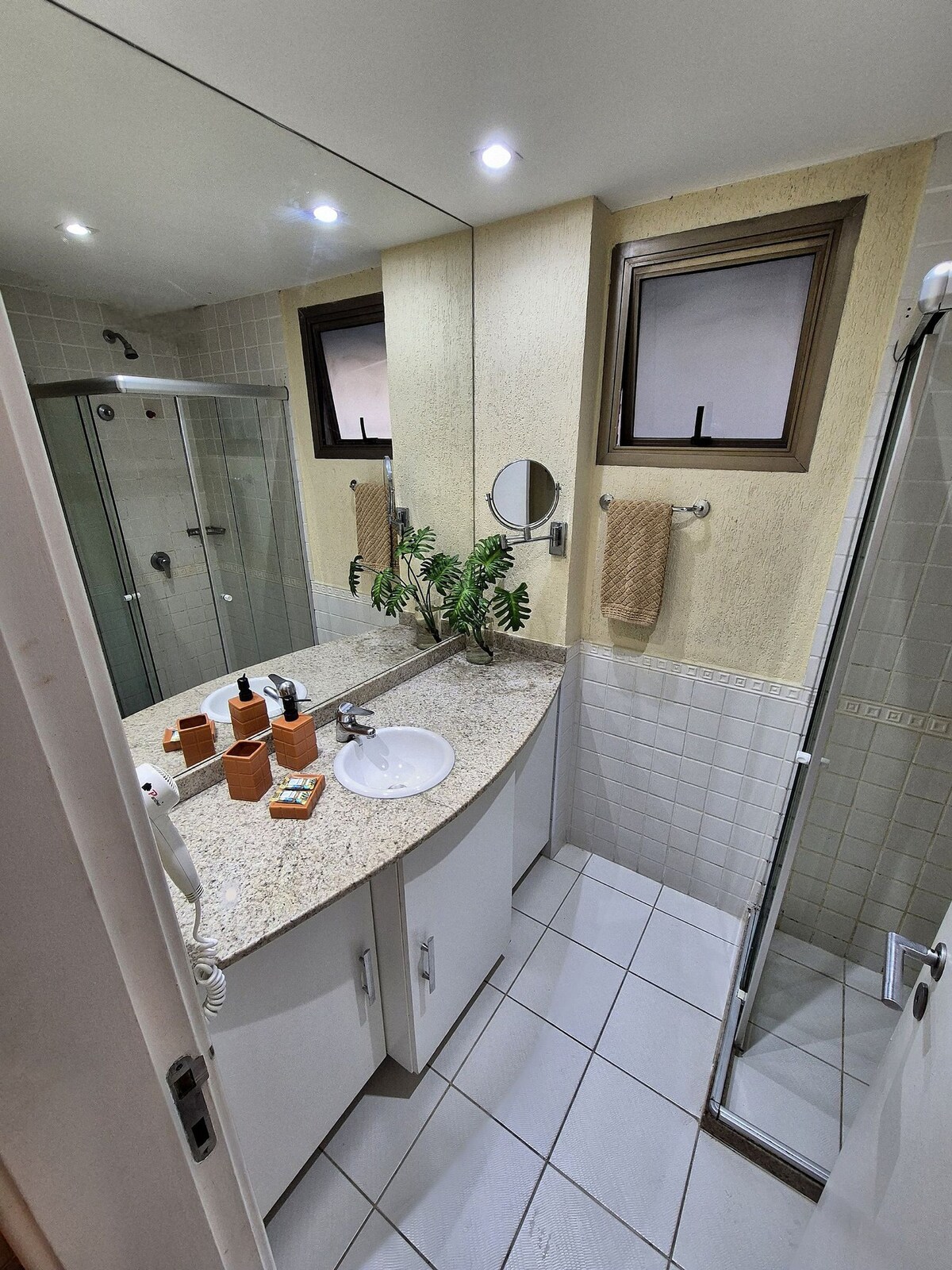 A clean bathroom space features a granite countertop with a round sink and toiletries arranged neatly. Two large mirrors reflect the area, providing ample lighting from ceiling fixtures. A glass shower stall is visible, along with leaf decor and hand towels hanging by the walls.