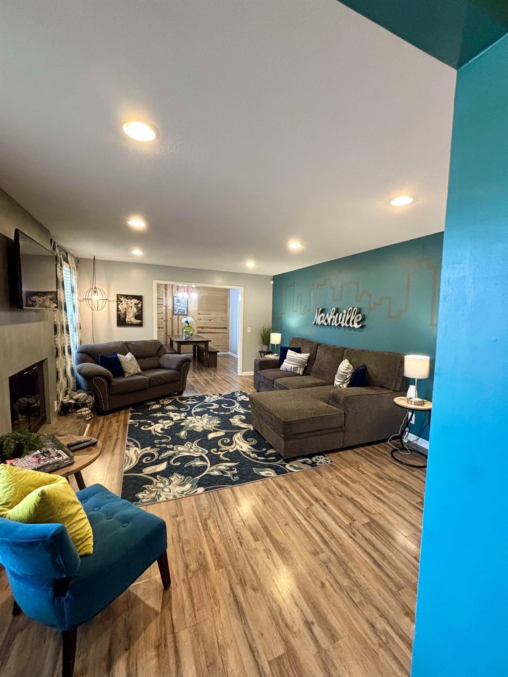 The living room features a mix of seating options, including two sofas and a chair, arranged around a centrally placed area rug. A wall displays the word 'Nashville' in a creative font, while light fixtures enhance the soft ambiance of the space.
