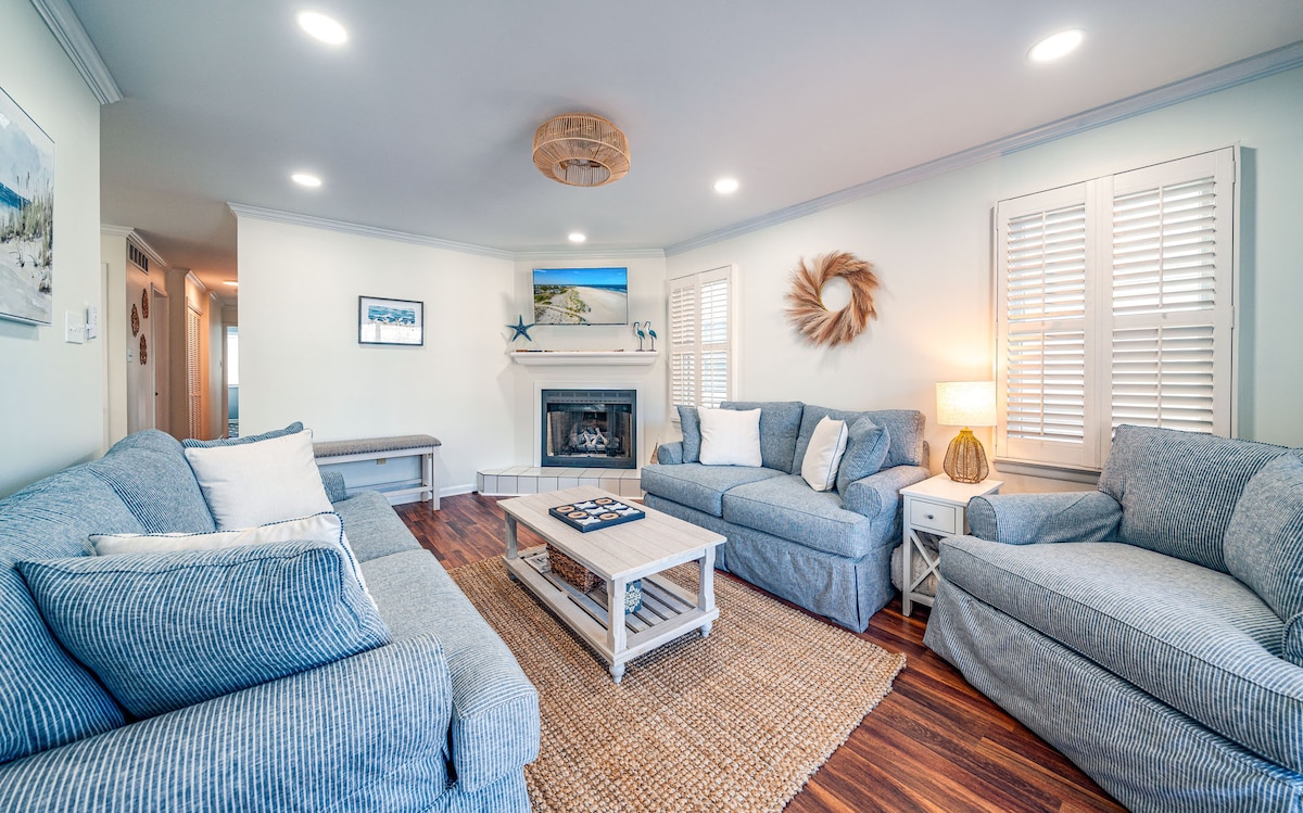 A bright living area features soft blue furniture arranged around a central coffee table. The space is complemented by a warm, neutral area rug and a decorative wreath on the wall. Natural light enters through the shuttered windows, creating a welcoming ambiance.